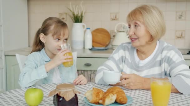 Χαριτωμένο κοριτσάκι πίνει χυμό και μιλάει με τη γιαγιά απολαμβάνοντας συνομιλία μιλώντας στην κουζίνα — Αρχείο Βίντεο