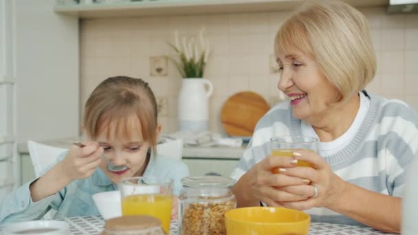おばあちゃんが笑顔で話している間に家で健康的な朝食を持っている愛らしい子供 — ストック動画