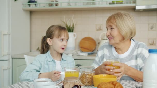 Медленное движение пожилой женщины, разговаривающей с внучкой, пока милая девушка завтракает на кухне — стоковое видео