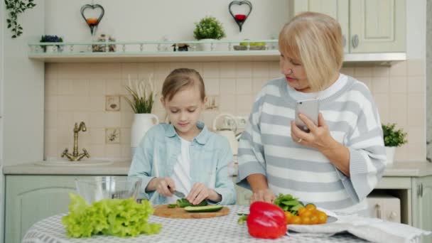Niña cortando verduras haciendo ensalada mientras la abuela con teléfono inteligente hablando niño enseñanza — Vídeos de Stock