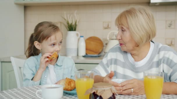 Eldrely woman and little girl having breakfast at home in kitchen talking laughing — Stock Video