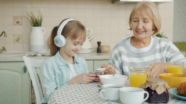 Повільний рух маленької дівчинки за допомогою смартфона і прослуховування музики в навушниках за кухонним столом з бабусею — стокове відео