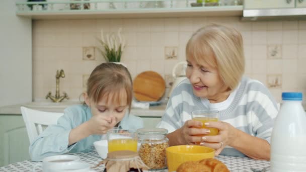 Little girl eating breakfast at home and talking to caring grandmother at table — Stock Video