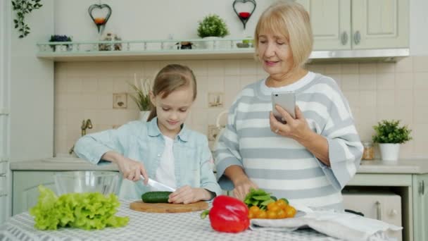 Abuela con smartphone enseñando a su nieta a cocinar ensalada en casa — Vídeo de stock