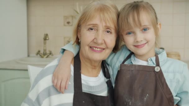 Retrato de niña y mujer madura con delantales de pie en la cocina abrazando y mirando a la cámara — Vídeo de stock