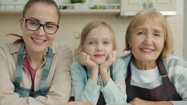 Ritratto di nonna, mamma e bambino sorridenti seduti in cucina indossando grembiuli guardando la macchina fotografica — Video Stock