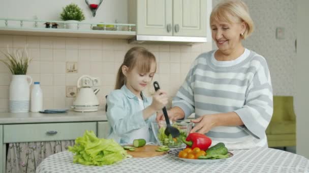 Niña cocinando ensalada mezclando verduras en un tazón y charlando con la alegre abuela en la cocina en casa — Vídeo de stock