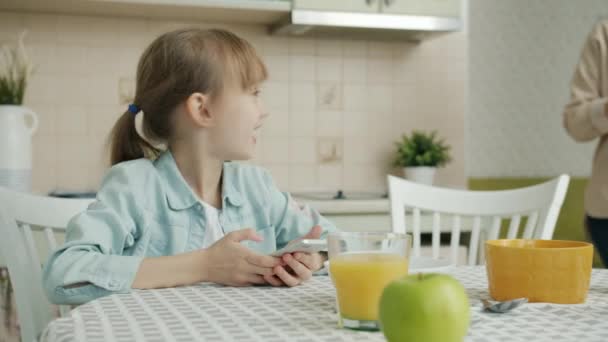 キッチンでスマートフォンを使っている幸せな子供に新鮮なクロワッサンをもたらす若い女性 — ストック動画