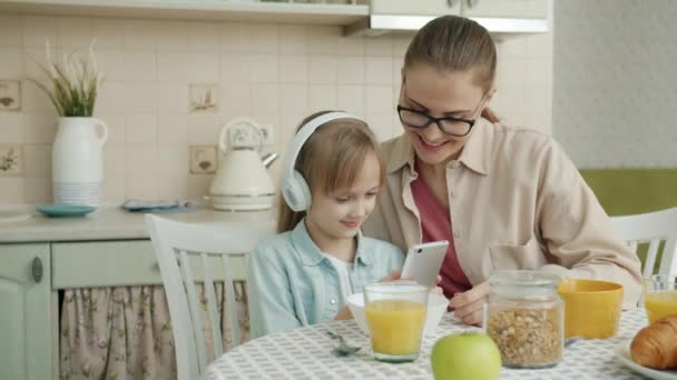 Медленное движение счастливой маленькой девочки и любящей матери при помощи смартфона и прослушивании музыки за кухонным столом — стоковое видео