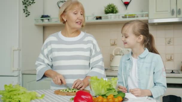 Anciana preparando ensalada y alimentando a su nieta con chuletas de pepino riéndose en casa — Vídeos de Stock