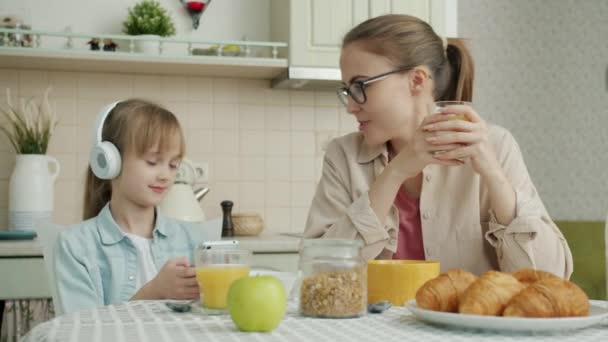 Mère et fille parlent à la table de cuisine pendant que la fille écoute de la musique et utilise un smartphone — Video