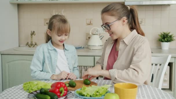 Молода жінка готує салат і годує милу маленьку доньку зі скибочками огірків на кухні — стокове відео