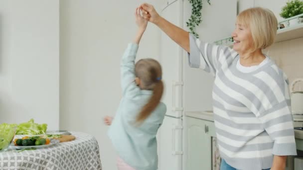 Радісна бабуся і мила дівчинка танцює на кухні вдома разом насолоджуючись музикою — стокове відео