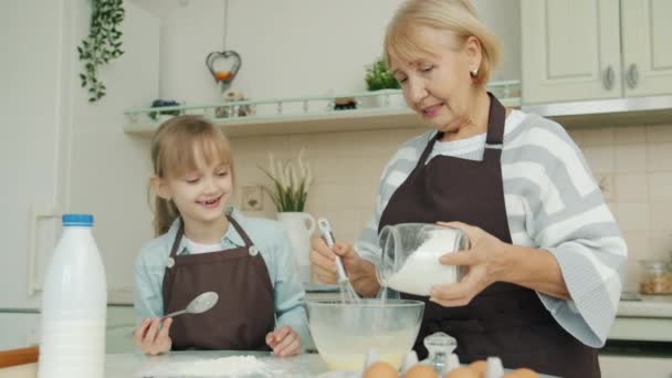 Femme âgée et petite fille dans des tabliers cuisine ajoutant de la farine à la pâte et parlant — Video