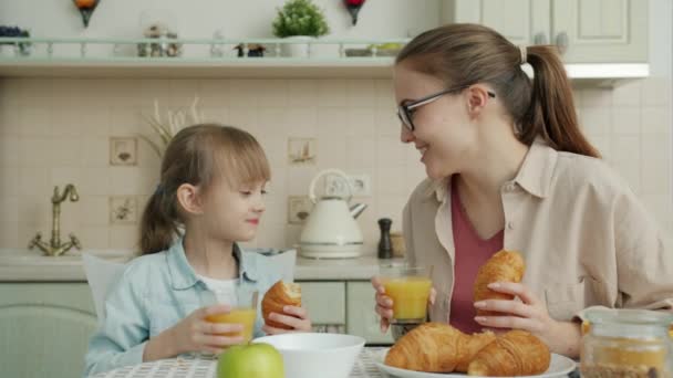 Молодая женщина и маленькая девочка едят круассаны и пьют сок разговаривая за завтраком — стоковое видео