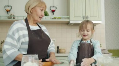 Yaşlı bir kadın hamur işi pişirirken küçük bir kız una bulanmış ellerini çırpıyor ve gülüyor.