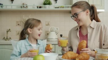 Neşeli çocuk ve neşeli anne modern evde kruvasan yiyor ve meyve suyu bardakları tıkırdıyor.