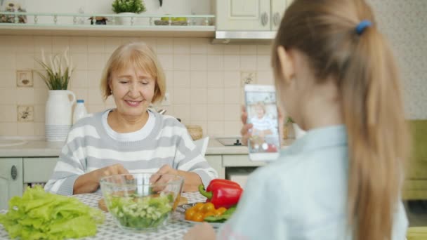 Alegre anciana posando para cámara de teléfono inteligente y ensalada de cocina mientras el niño toma la foto — Vídeos de Stock