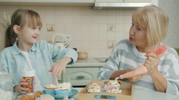 Äldre kvinna och ung flicka dekorera muffins hemma pratar njuta av kulinarisk aktivitet — Stockvideo