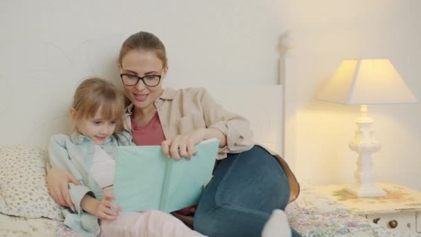 Заботливая мать читает книгу очаровательной маленькой девочке, сидящей в постели вместе — стоковое видео