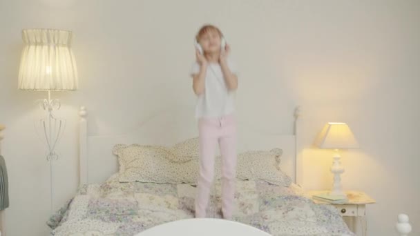 Joyful little girl jumping dancing in bed listening to music through headphones in bedroom — Stock Video