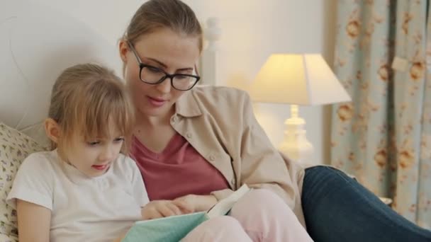 Молода жінка читає книгу маленькій дівчинці, яка розслабляється в ліжку в сучасній квартирі — стокове відео