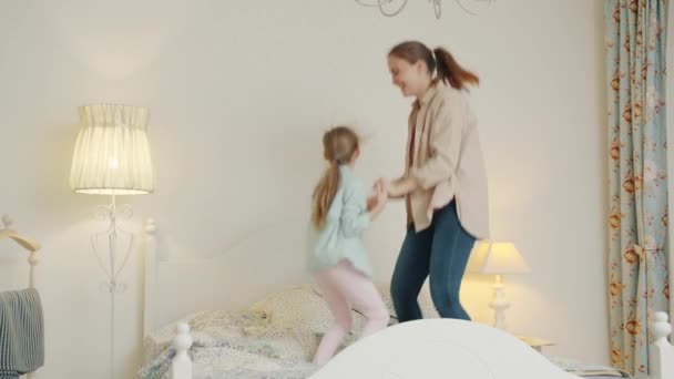 어린 소녀와 즐거운 어머니가 침대에서 손을 잡고 뛰면서 집에서 함께 즐거운 시간을 보내고 있다 — 비디오