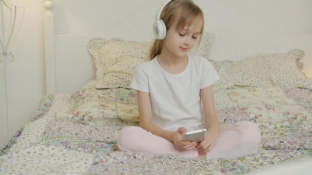 Fröhliches Kind genießt Musik im Kopfhörer und berührt Smartphone-Bildschirm im Bett — Stockvideo