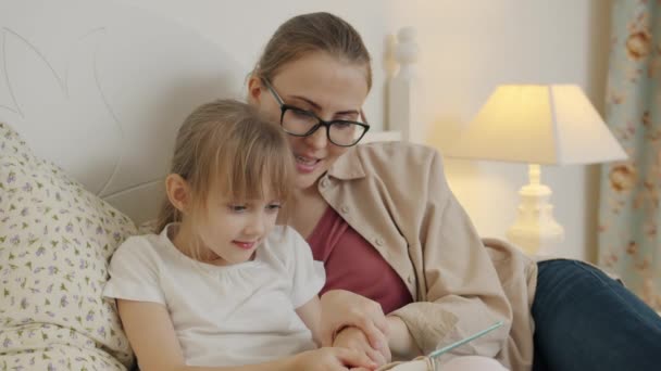 Маленька дівчинка вчиться читати книгу з матір'ю в ліжку, насолоджуючись літературою — стокове відео