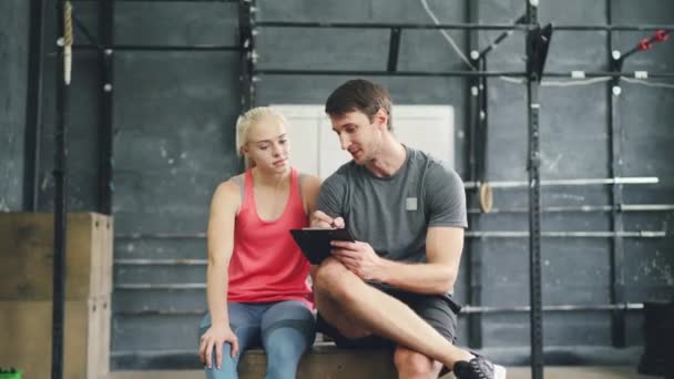 Radosna dziewczyna i facet rozmawiający relaks po treningu w crosfit siłowni, trener pisze w wykresie — Wideo stockowe