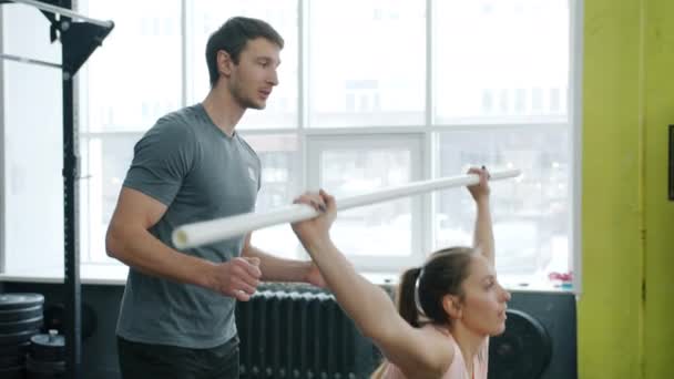 Odchylona młoda kobieta kuca z ciężkim barem ćwicząc na siłowni z instruktorem — Wideo stockowe
