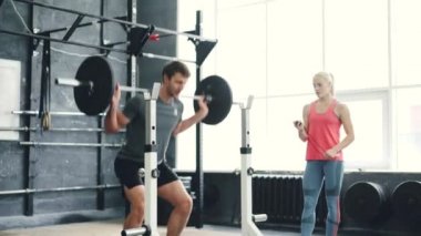 Spor giyimli bir adam spor salonunda halterli bir şekilde kadın koçla antrenman yapıyor.