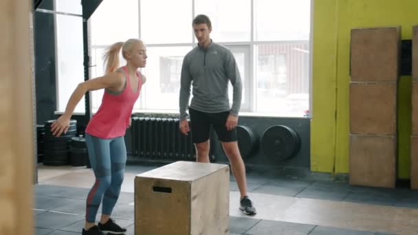 Chica en ropa deportiva saltando en el entrenamiento de caja mientras entrenador crossfit viendo — Vídeos de Stock