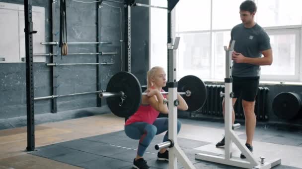 Słodkie blondynka squatting z sztangą wewnątrz działa z profesjonalnym trenerem — Wideo stockowe