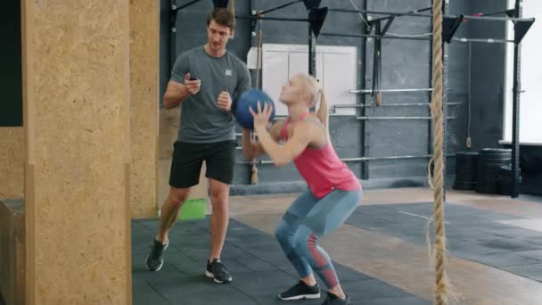 Mouvement lent de la formation cherful fille dans la salle de gym avec entraîneur jetant balle lourde squatting — Video