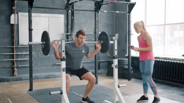 Сильний молодий чоловік присідає з важким барбелом, який працює з інструктором у спортзалі — стокове відео