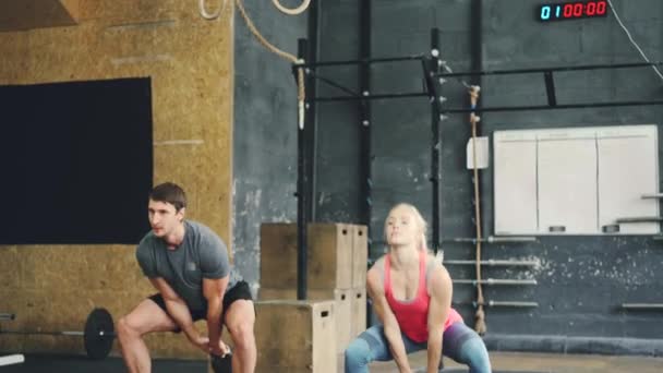 Lány és a srác a sport felszerelés guggoló kettlebells alatt crossfit edzés tornaterem — Stock videók
