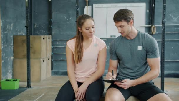 Coach en jonge vrouw praten ontspannen in de sportschool, man is het schrijven van onderwijs meisje — Stockvideo