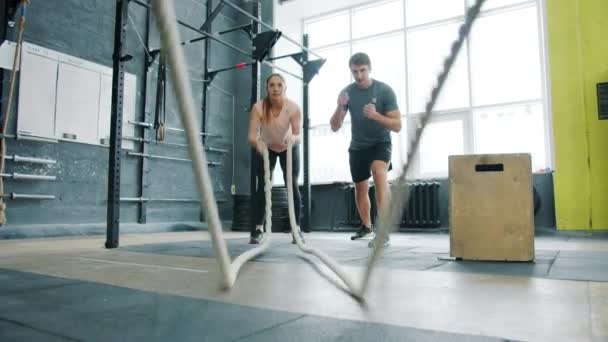 Девушка тренируется с боевыми веревками в кроссфит-тренажерном зале с профессиональным тренером молодым человеком — стоковое видео