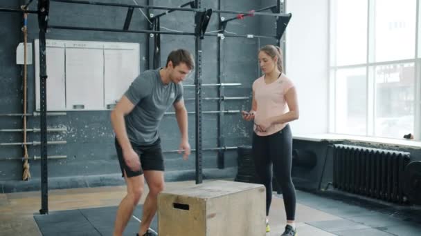 Mladý silný muž skákání na dřevěné krabici cvičení s crossfit trenér v tělocvičně — Stock video