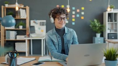Neşeli ofis çalışanı dizüstü bilgisayar kullanıyor kulaklıkla müzik dinliyor.