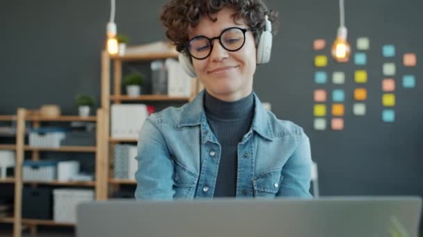 Uśmiechnięta bizneswoman nosząca słuchawki pracująca z laptopem w biurze ciesząca się muzyką — Wideo stockowe