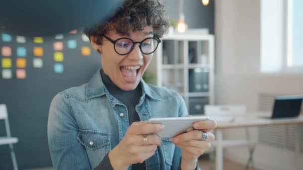 Веселая девушка расслабляется в офисе, играя в видеоигры касаясь экрана смартфона — стоковое видео