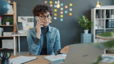 Neşeli ofis çalışanı cep telefonuyla konuşuyor ve modern işyerindeki laptop ekranına bakıyor.