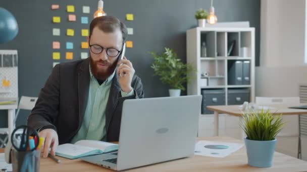 Chico alegre que trabaja en la oficina haciendo la llamada del teléfono móvil y usando el ordenador portátil que toma notas en el cuaderno — Vídeo de stock