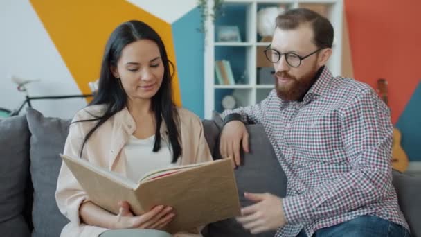 Pareja feliz viendo fotos en el álbum y hablando relajante en el sofá en casa — Vídeos de Stock