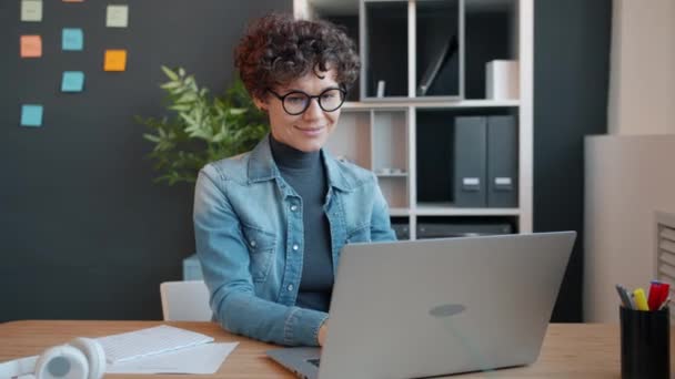 Веселая девушка занята работой с ноутбуком в офисе, печатая и глядя на экран улыбаясь — стоковое видео