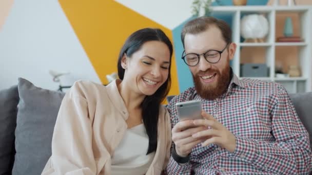 Marido e esposa usando smartphone rindo sentado no sofá em casa juntos — Vídeo de Stock