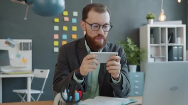 Modern ofiste akıllı telefondan mobil video oyunu oynayan mutlu genç adam.