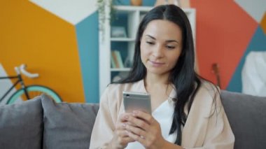 Akıllı telefonu tutan çekici genç bir kadın ekrana dokunuyor ve gülümsüyor evde dinleniyor.